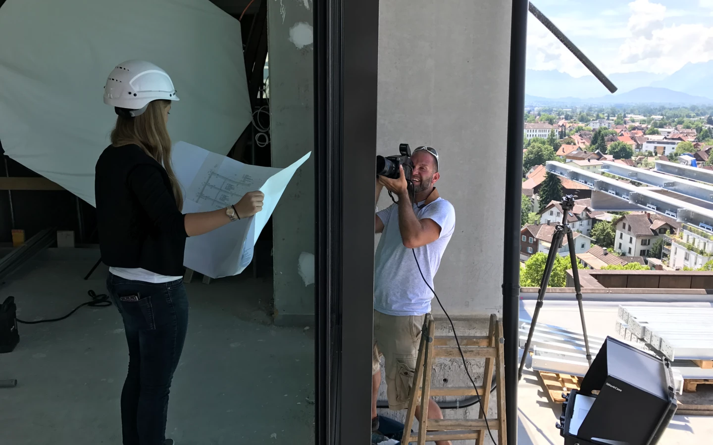 Eine Frau mit Schutzhelm und Bauplänen sowie ein Fotograf auf einer Leiter mit Kamera bei einem Fotoshooting auf einer Baustelle.