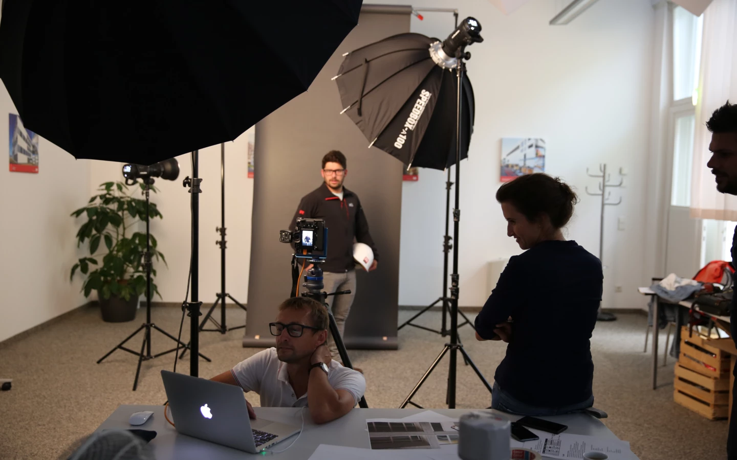 Männer und Frauen bei einem Fotoshooting, ein Mann sitzt vor einem Laptop, während ein anderer Mann posiert und eine Frau zusieht.