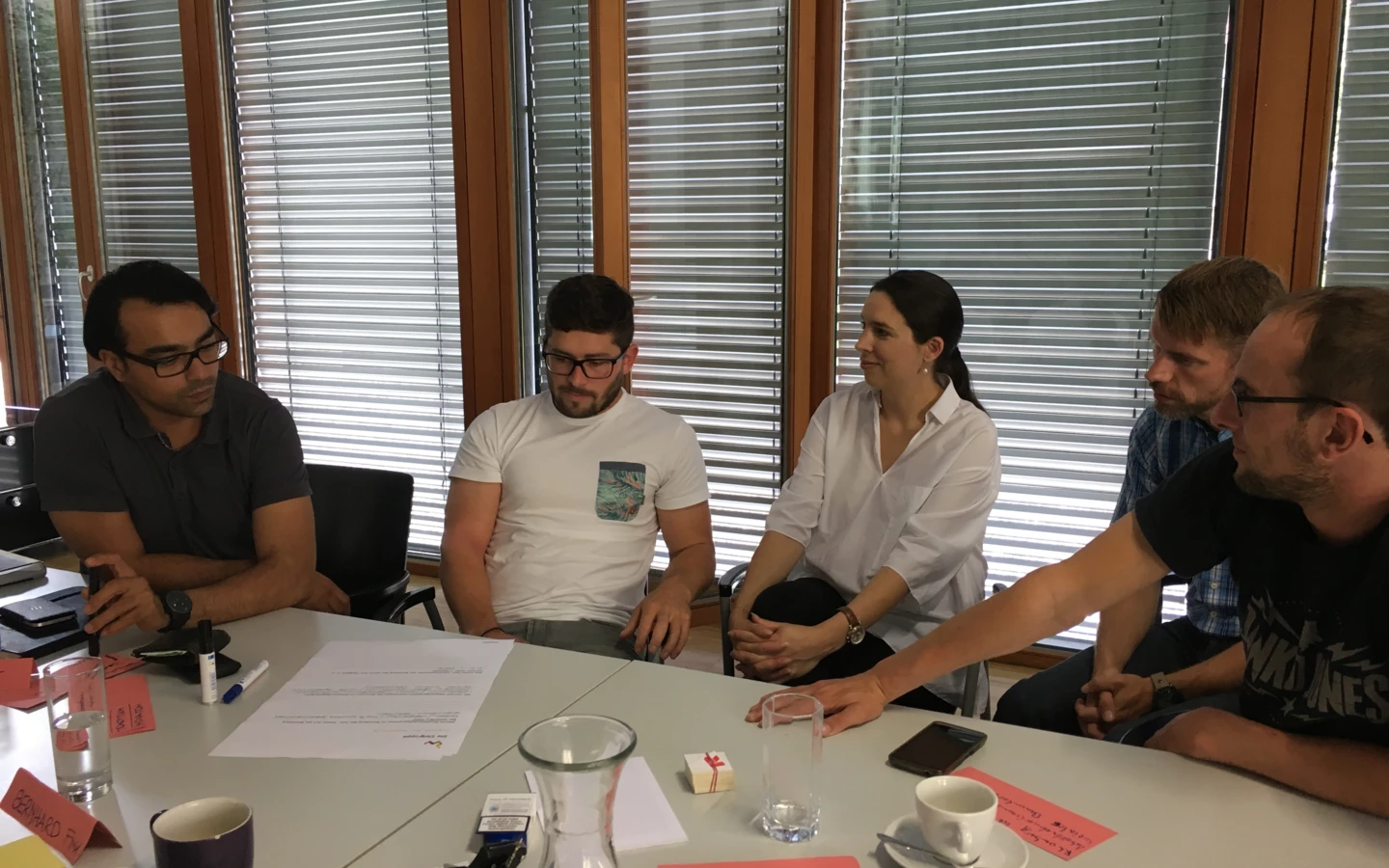 Fünf Personen sitzen an einem Tisch, besprechen Dokumente und trinken Kaffee in einem Workshop-Umfeld bei Rhomberg Bau.