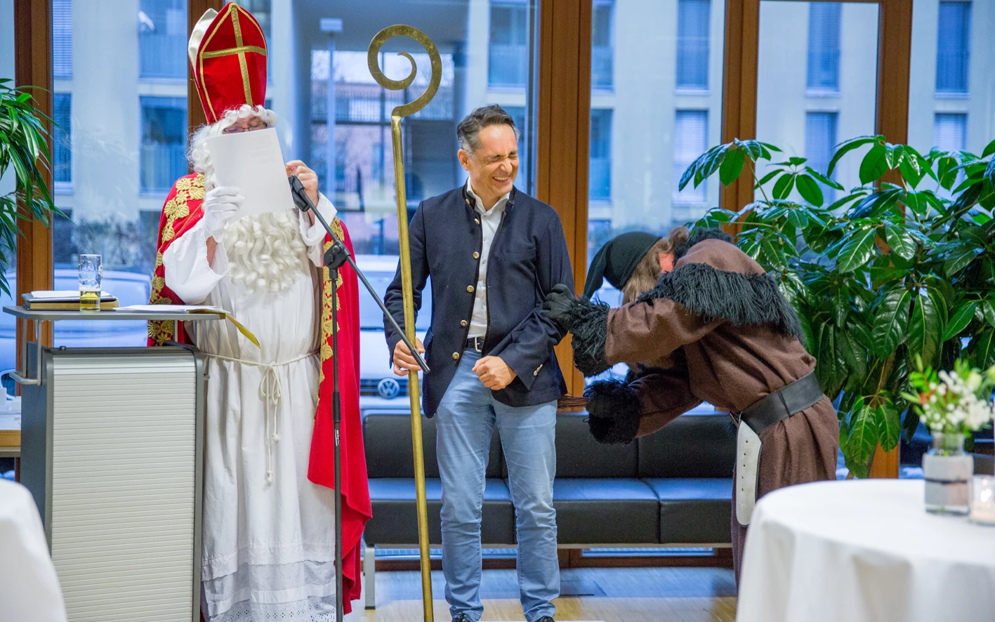 Ein Mann im Nikolaus-Kostüm liest von einem Blatt ab, während ein anderer Mann lachend einen Bischofsstab hält und ein Krampus zu seinen Füßen kauert (Hubert, Geburtstag).