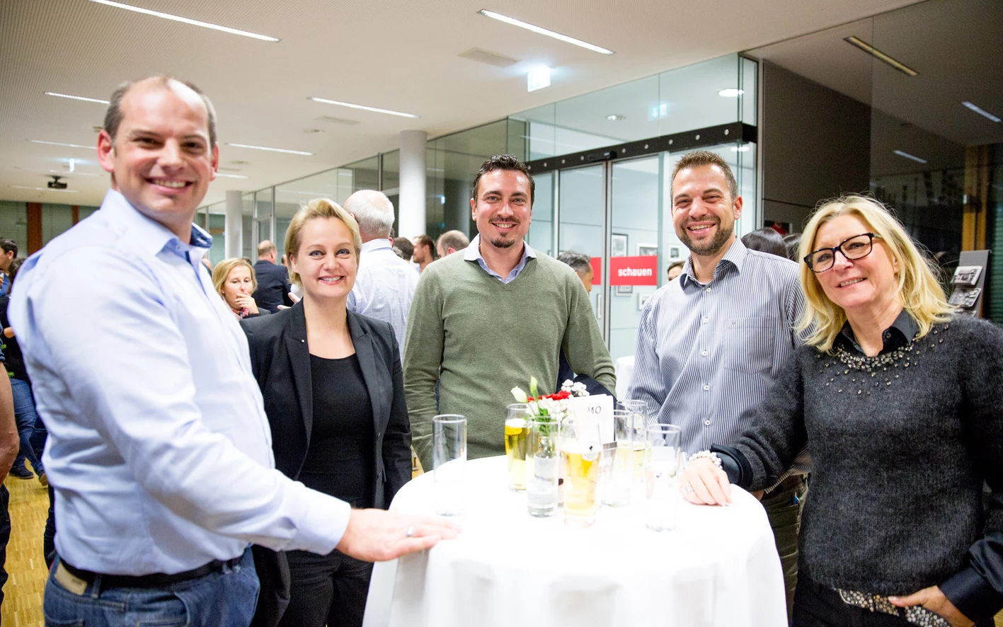 Fünf lächelnde Personen stehen um einen runden Tisch mit Getränken und Blumen bei einer Veranstaltung (Hubert, Geburtstag)