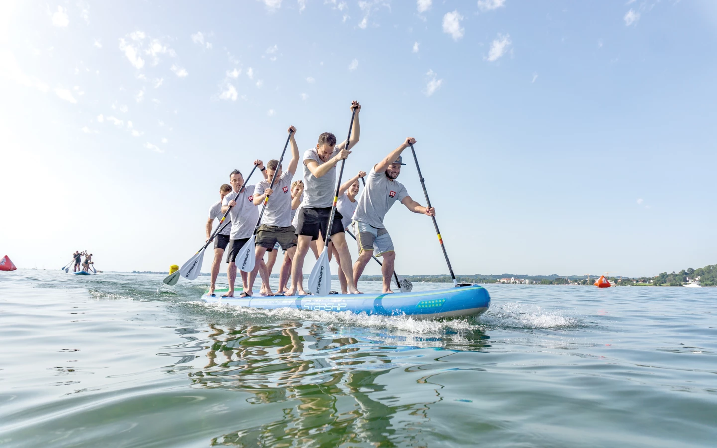 Eine Gruppe von sechs Personen paddelt gemeinsam stehend auf einem großen blauen SUP-Board über klares Wasser unter strahlend blauem Himmel (MathiasJoschika).