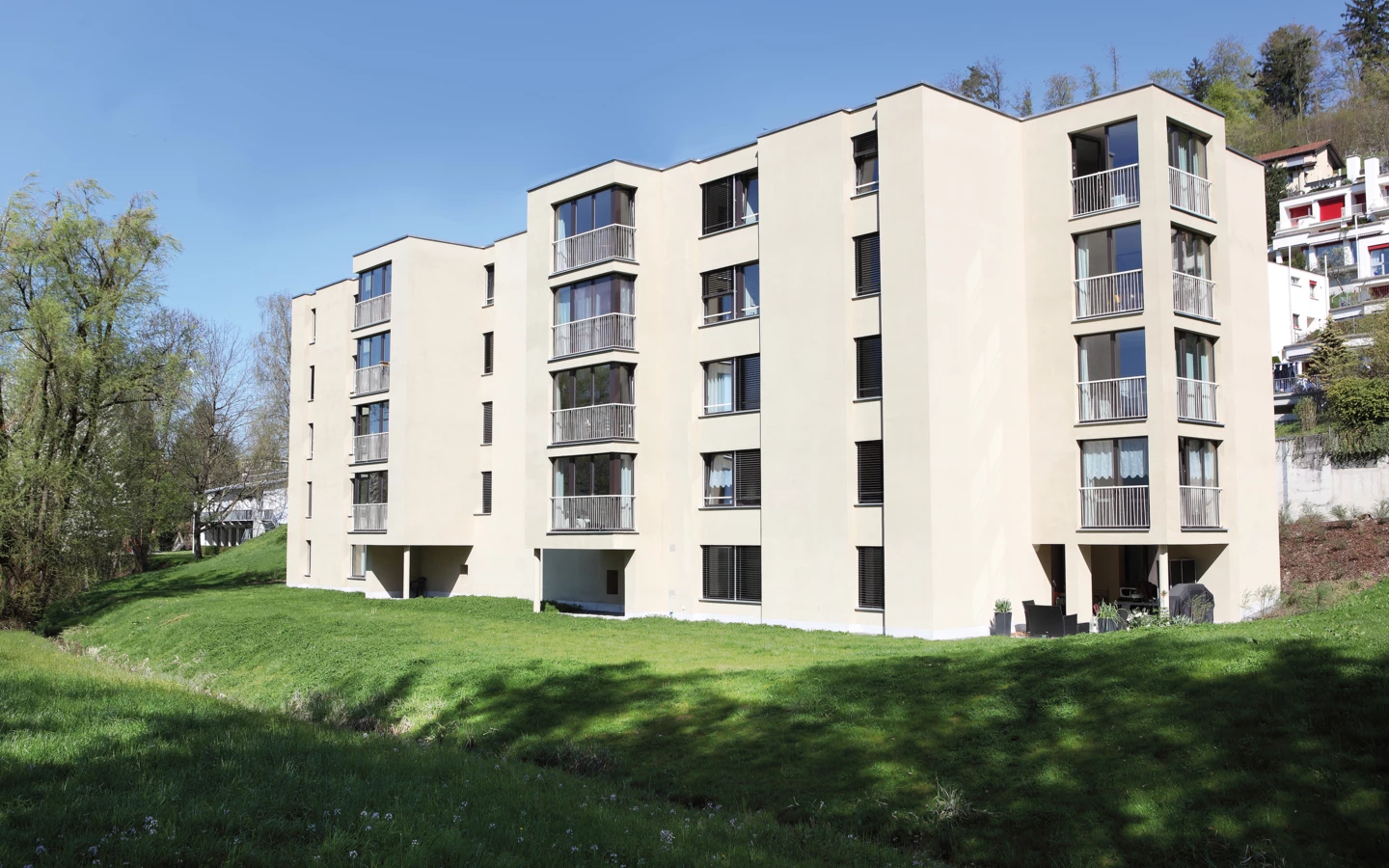 Modernes beige Mehrfamilienhaus mit Balkonen und Jalousien, umgeben von grünen Wiesen und Bäumen unter blauem Himmel