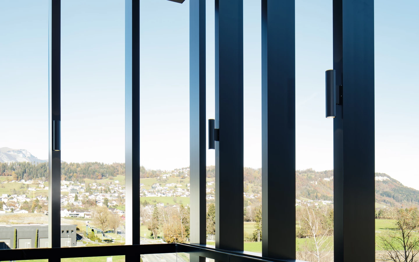 Ein moderner Raum mit großen Fenstern, die den Blick auf ein grünes Tal mit Häusern und Bergen freigeben. Schwarze Fliesen auf dem Boden (Edenta).