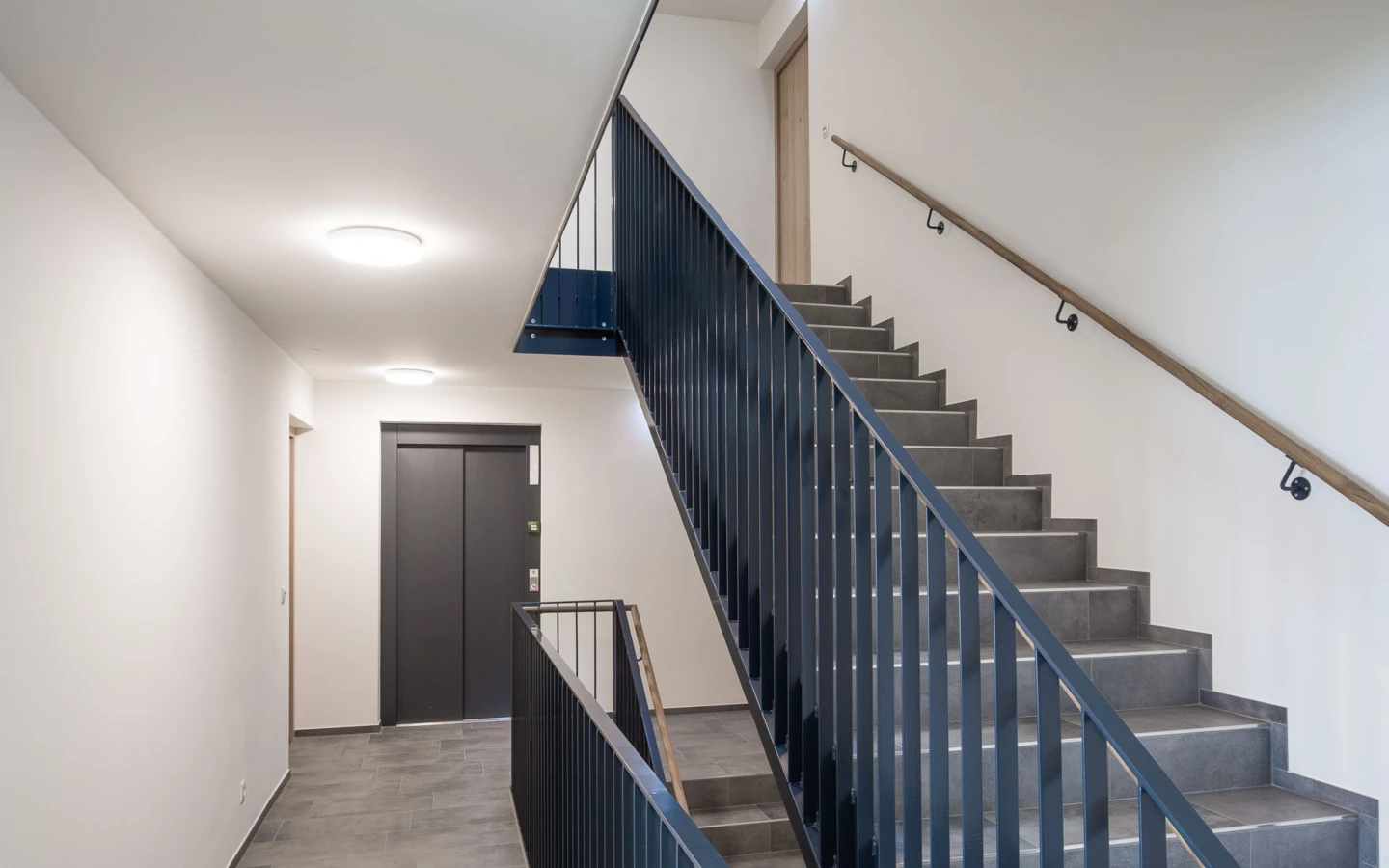 Modernes Treppenhaus mit grauem Fliesenboden, dunklem Metallgeländer, hellem Holzhandlauf und einem Aufzug in der Wohnanlage Bahnhofsstrasse Vandans (Stiegenhaus).