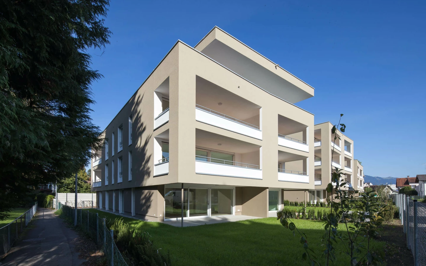 Moderne Wohnanlage Widum Lustenau mit beigefarbenen Fassaden und Balkonen, umgeben von Grünflächen und einem Fußweg unter klarem Himmel.