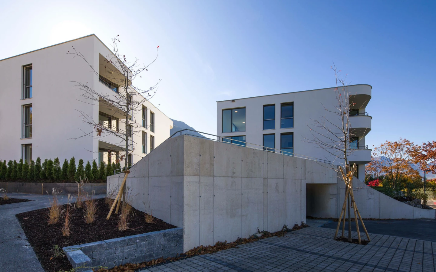 Wohnanlage Wolf-Huber-Straße in Feldkirch mit weißen Gebäuden, Tiefgarageneinfahrt, begrünten Beeten und gepflastertem Gehweg an einem sonnigen Tag.