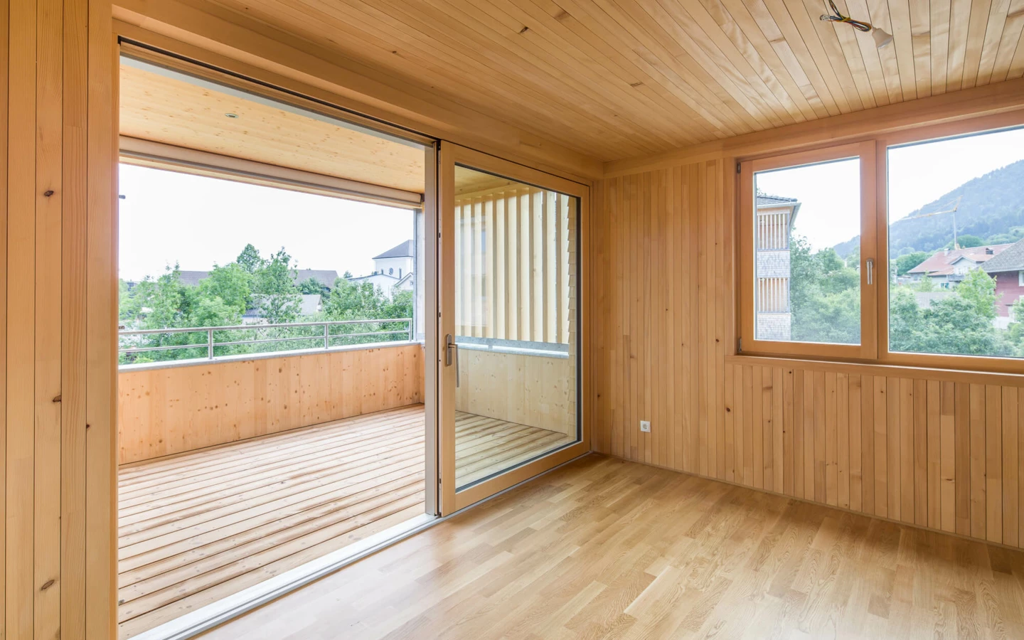 Ein Zimmer mit hellen Holzpaneelen an Wänden und Decke, Holzboden, Schiebetür zum Balkon und Fenster mit Blick ins Grüne (Wohnanlage, Alberschwende).