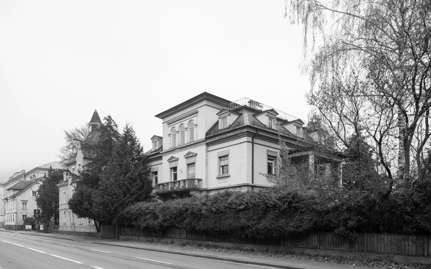 Schwarz-Weiß-Ansicht der Villa Reichsstraße, einer historischen Residenz mit großem Balkon hinter einer hohen Hecke, an einer Straße in Bregenz.