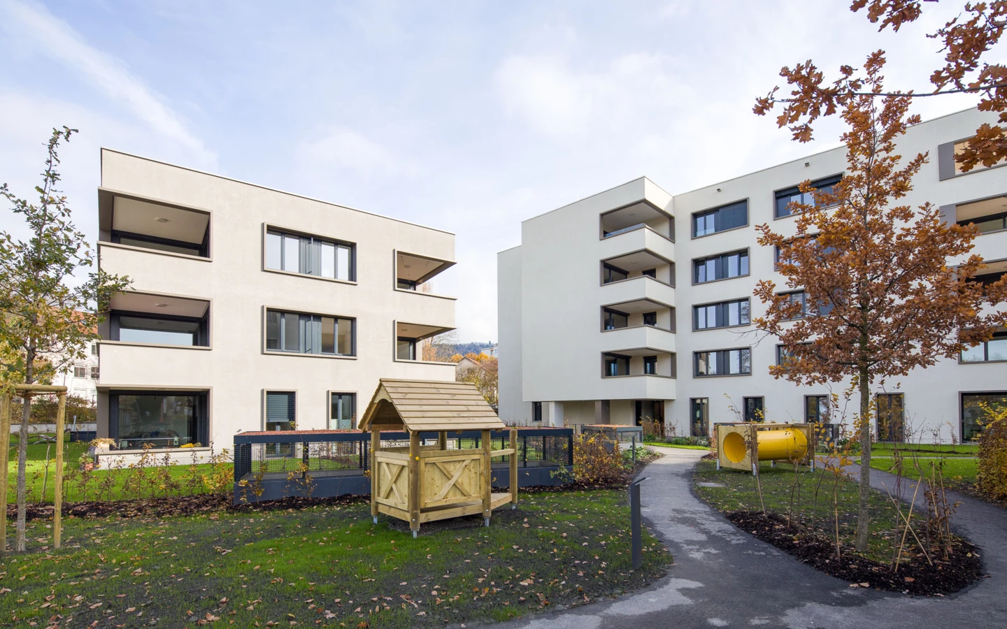 Zwei moderne weiße Wohngebäude mit Balkonen, umgeben von einem begrünten Hof mit Spielhaus und Tunnel (Bäumleareal)