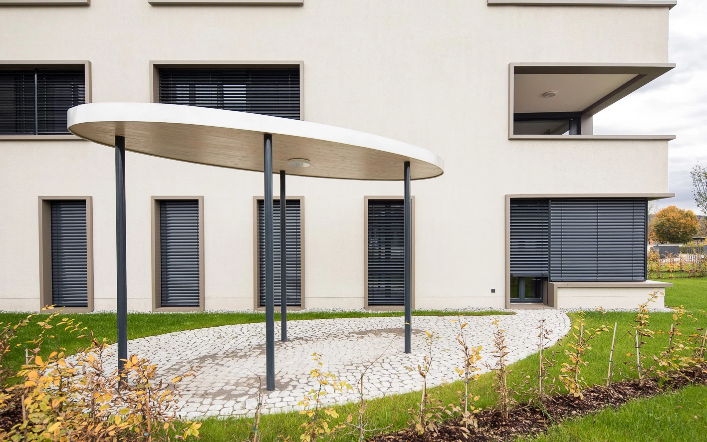 Eingangsbereich eines modernen Gebäudes mit ovalem Vordach, gepflastertem Weg und Grünflächen mit jungen Sträuchern (Baeumlequartier).