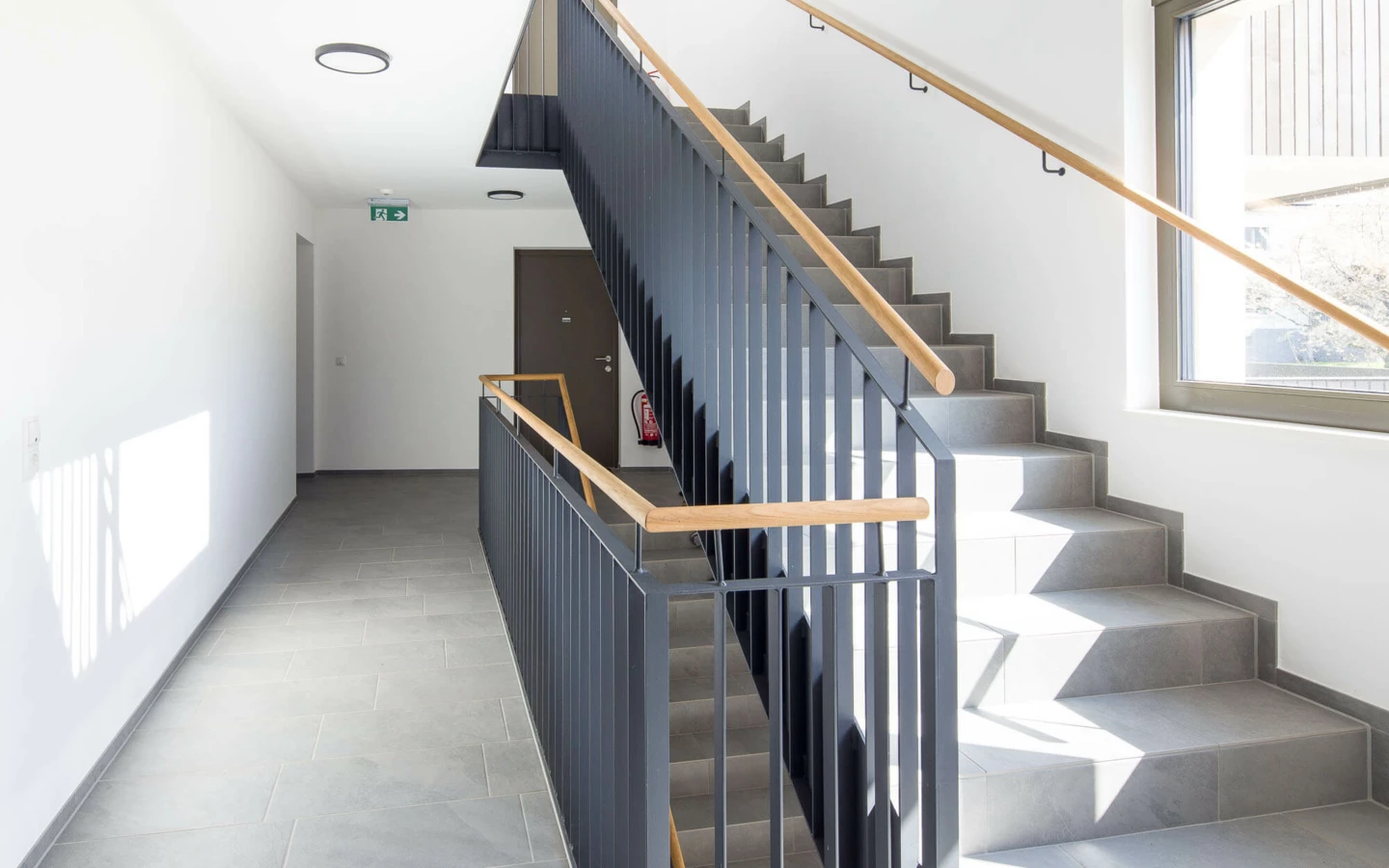 Modernes Treppenhaus mit grauen Fliesen, Metallgeländer und Holzhandläufen in der Wohnanlage Feldgasse in Dornbirn.