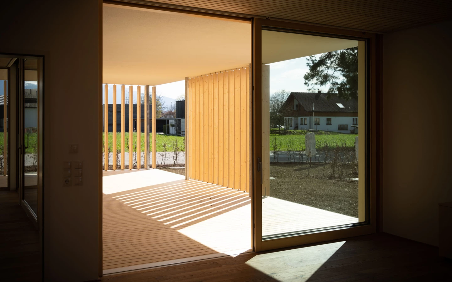 Ein moderner, offener Holzpavillon mit Lamellen und Holzboden verbindet Innen- und Außenbereich, unterstrichen durch Sonnenlichtstreifen (Wolfurt, Dieter).