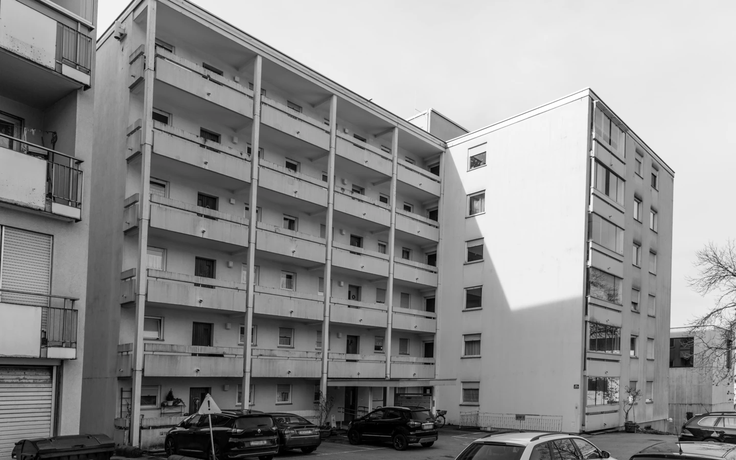 Eine Schwarz-Weiß-Aufnahme eines hohen Wohnhauses mit vielen Balkonen und geparkten Autos davor (Römerstraße).