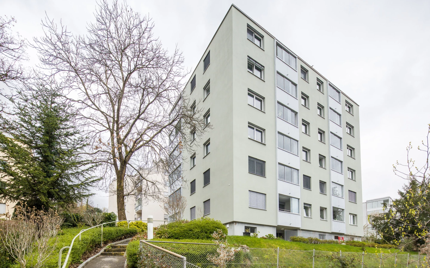 Mehrgeschossiges hellgrünes Gebäude mit vielen Fenstern und Balkonen, umgeben von Bäumen und einer Grünfläche mit einem geschwungenen Weg an einem bewölkten Tag (Römerstraße).