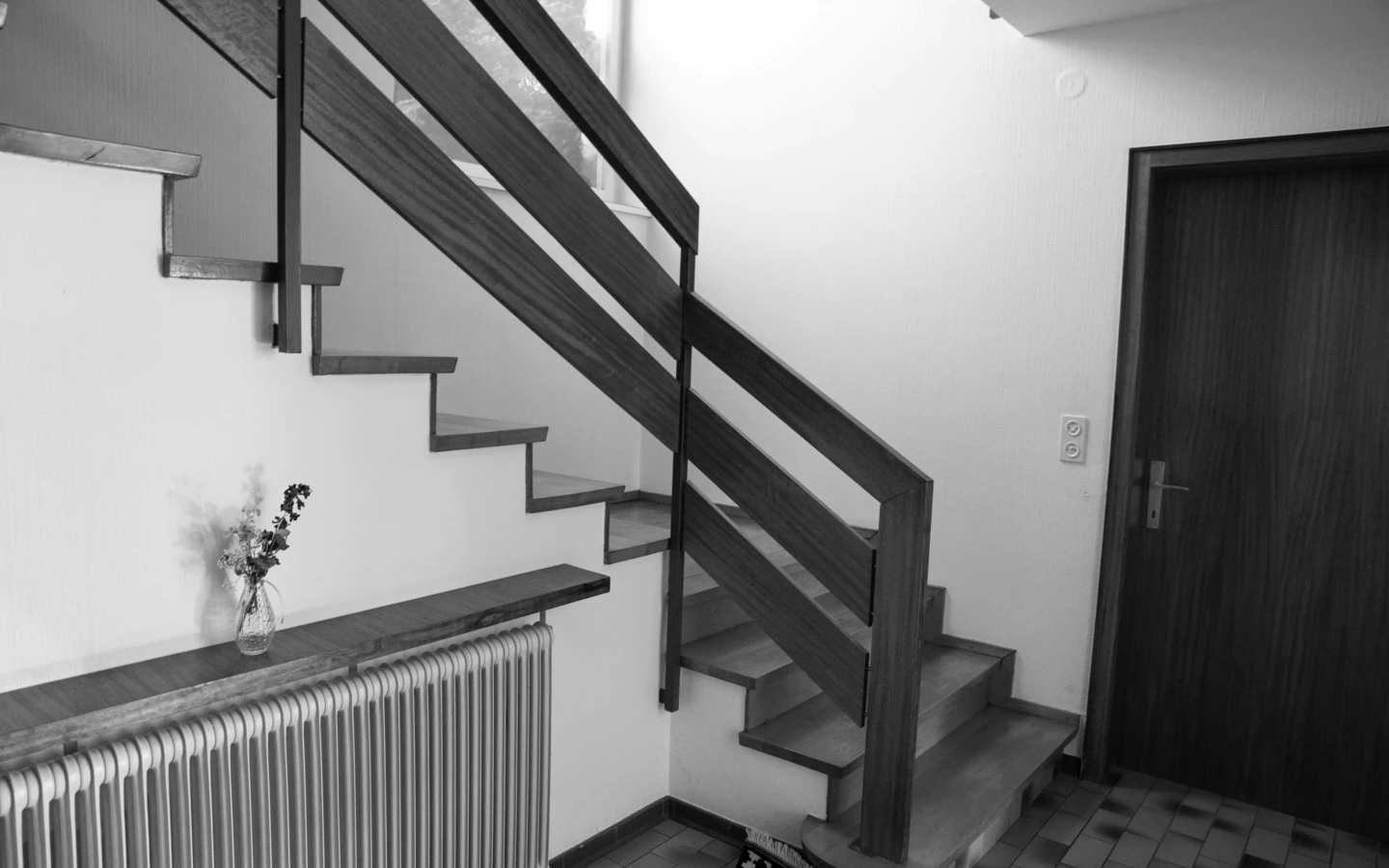 Schwarz-Weiß-Bild einer Treppe mit Holzstufen und Geländer, neben einem Heizkörper und einer Tür in einem Einfamilienhaus in Dornbirn (Generalsanierung, Holzoptik).