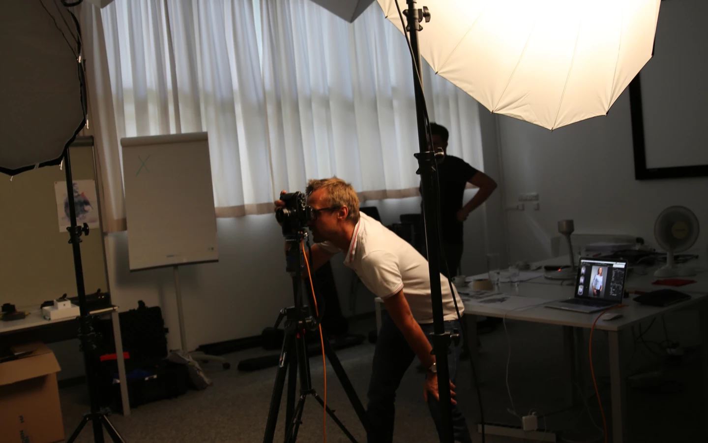 Ein Fotograf mit Brille fotografiert im Studio, umgeben von zwei Softboxen und einem Laptop mit Bildvorschauen.