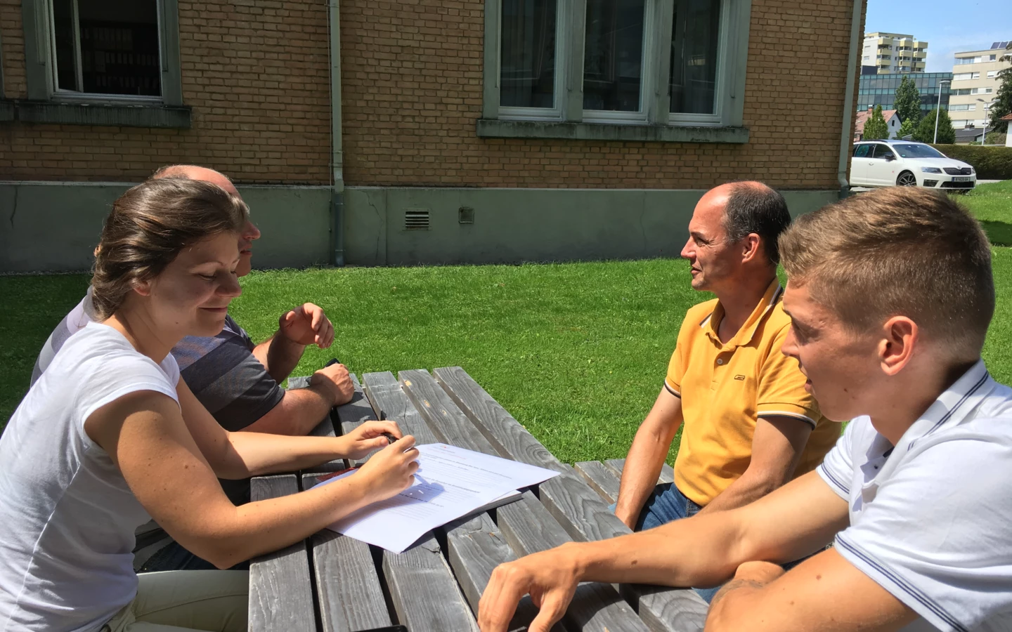 Vier Personen sitzen bei sonnigem Wetter an einem Holztisch im Freien, unterhalten sich und lesen Dokumente (Workshop).