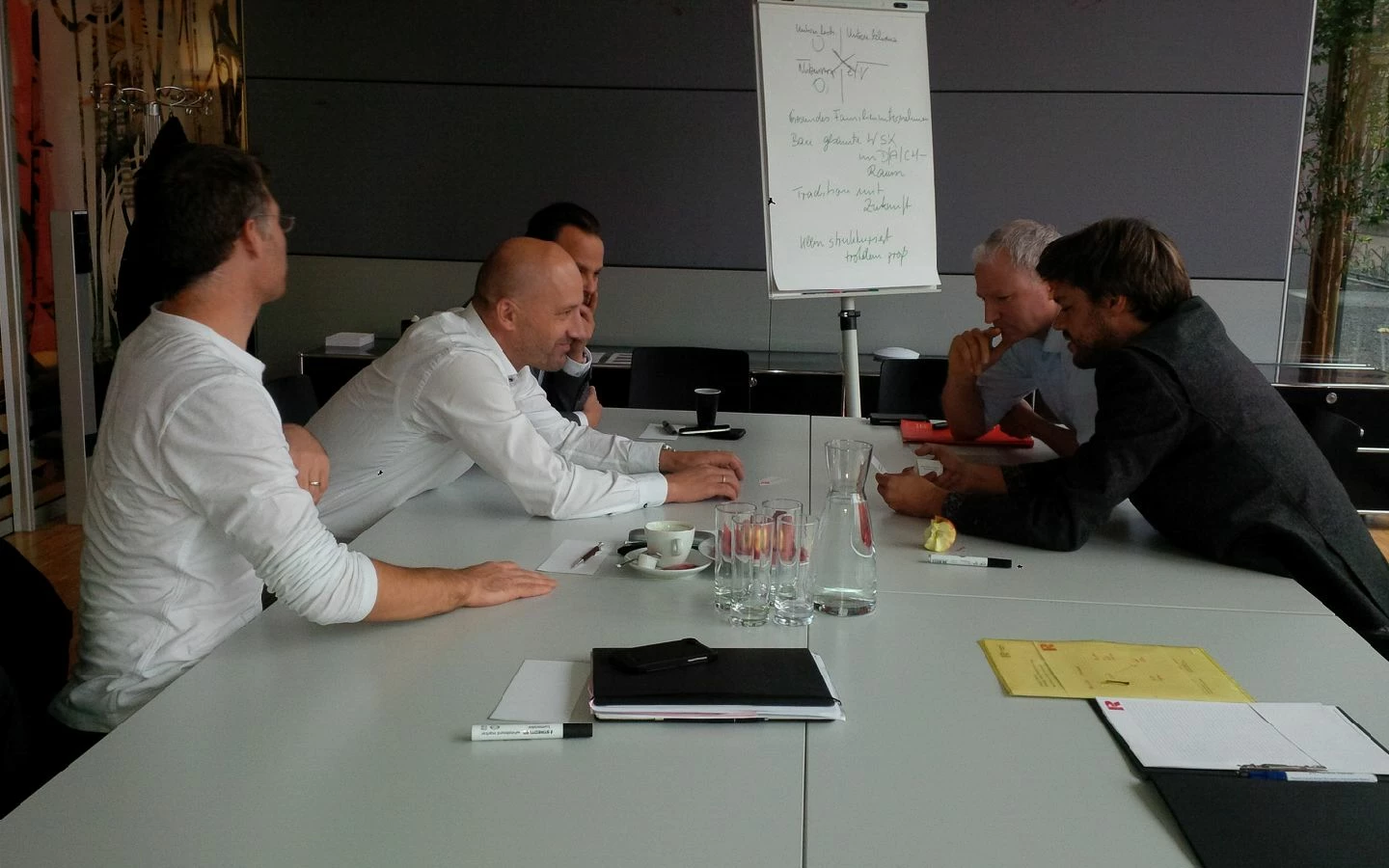 Fünf Männer sitzen an einem Konferenztisch mit Unterlagen und einem Flipchart im Hintergrund, bei einem Workshop (Führungskräfte).