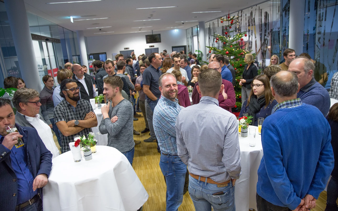 Eine große Gruppe von Menschen unterhält sich informell in einem hellen, modernen Raum mit einem Weihnachtsbaum, Stehtischen und Getränken (Hubert, Geburtstag).