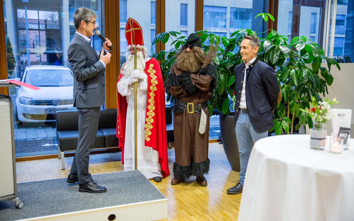 Ein Mann im Anzug spricht ins Mikrofon neben einem als Nikolaus verkleideten Mann, einem Mann in dunkler Kleidung mit Bart und einem Mann im Trachtenjanker (Hubert, Geburtstag).