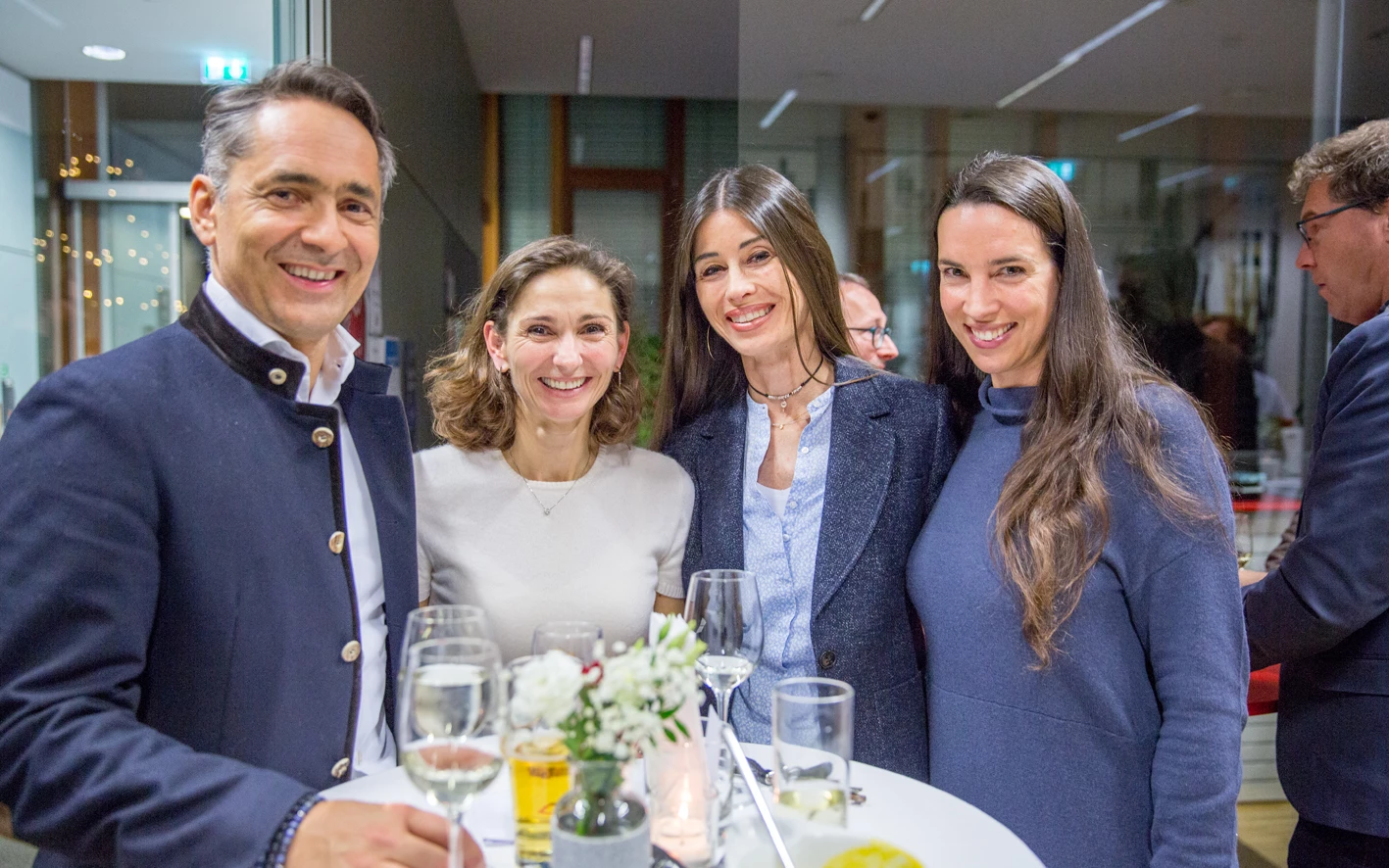 Vier lachende Personen, drei Frauen und ein Mann in Trachtenjacke, stehen an einem Stehtisch mit Getränken bei einer Veranstaltung (Hubert, Geburtstag).