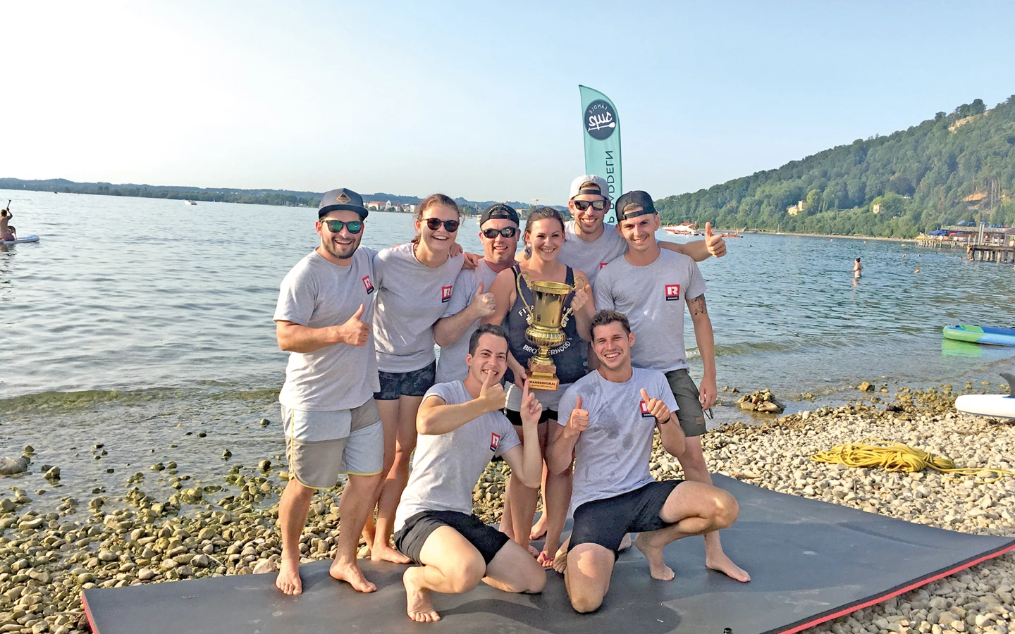 Eine Gruppe fröhlicher Kollegen in Shirts mit "R"-Logo und Sonnenhüten, die mit der gemeinsamen Trophäe am Seeufer für ein Foto posieren.