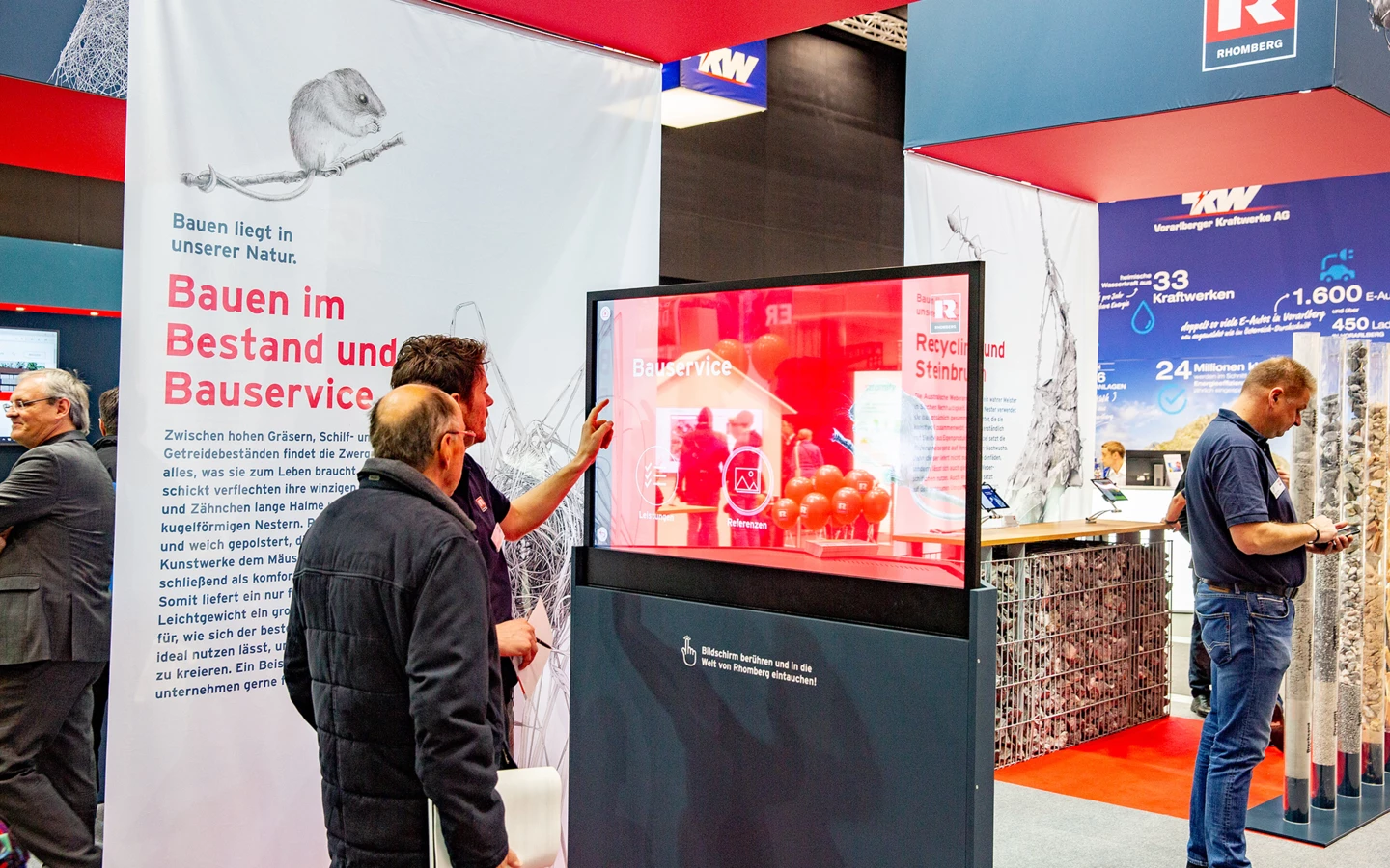 Besucher interagieren mit einem Touchscreen-Display am Rhomberg Bau Messestand, der "Bauservice" und "Recycling und Steinbruch" präsentiert.
