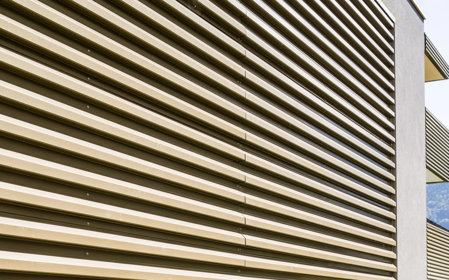 Seitenansicht einer modernen beige-braunen Fassade mit horizontalen Lamellen und vertikalen betongrauen Elementen. Teil der Wohnanlage Bahnhofsstrasse Vandans.