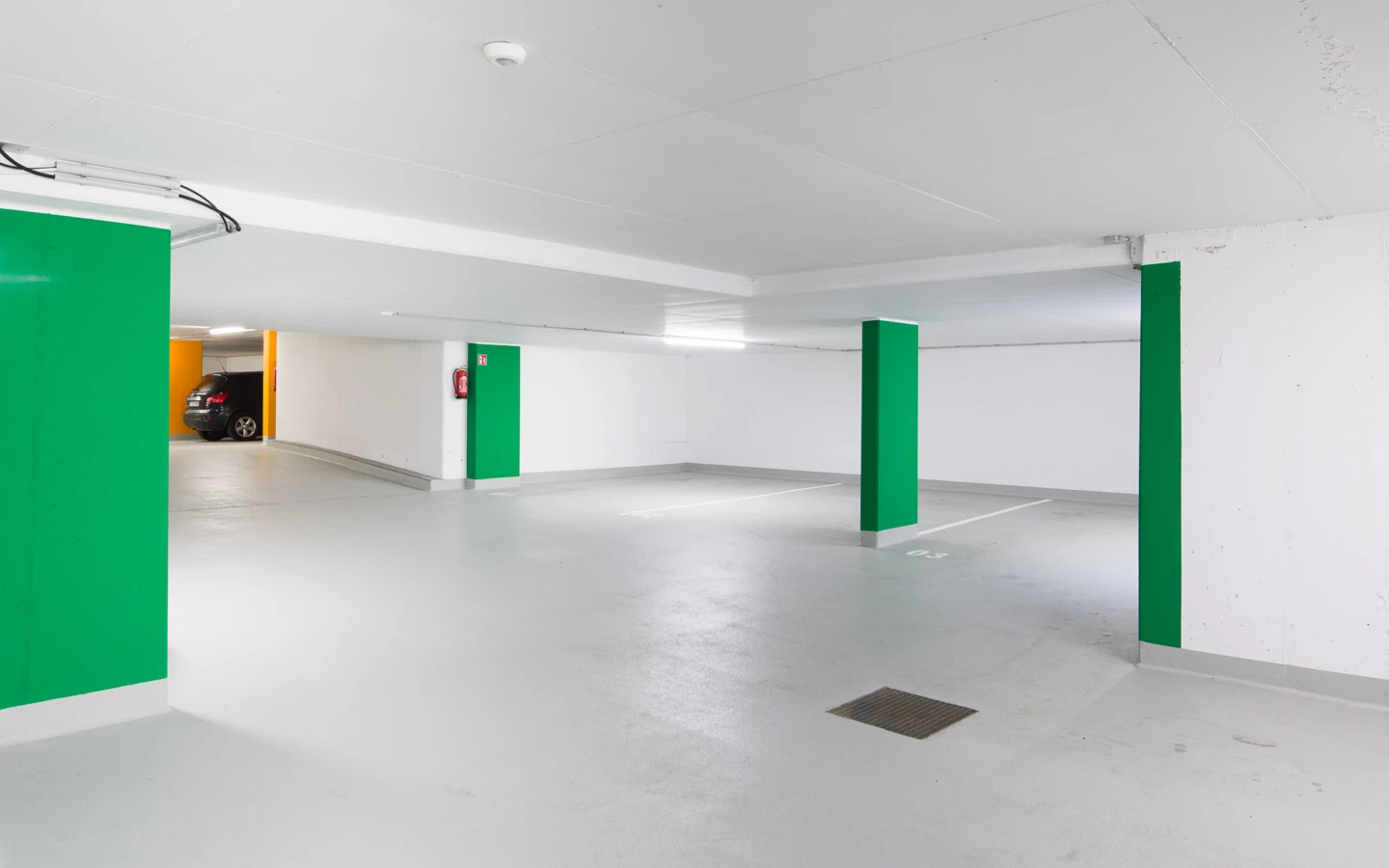 Eine leere, helle Tiefgarage mit grünen Säulen und einem schwarzen Auto am Eingang. Ein Feuerlöscher ist an der Wand sichtbar (Bahnhofstraße, Wohnanlage, Bahnhofsstrasse, Vandans).
