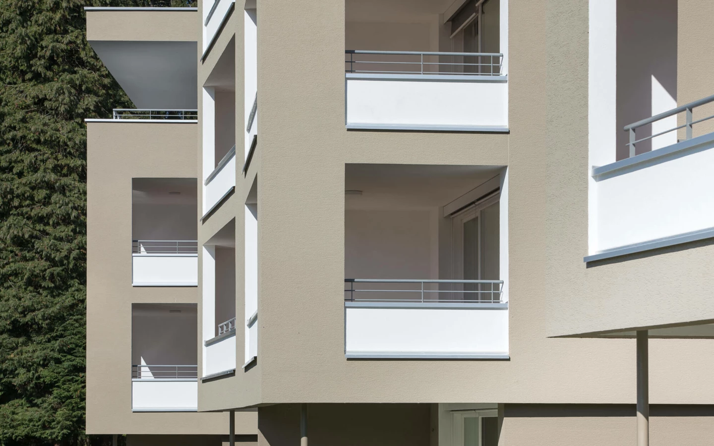 Detailansicht der sandfarbenen Fassade der Wohnanlage Widum Lustenau mit modernen Balkonen und hohen Bäumen im Hintergrund.
