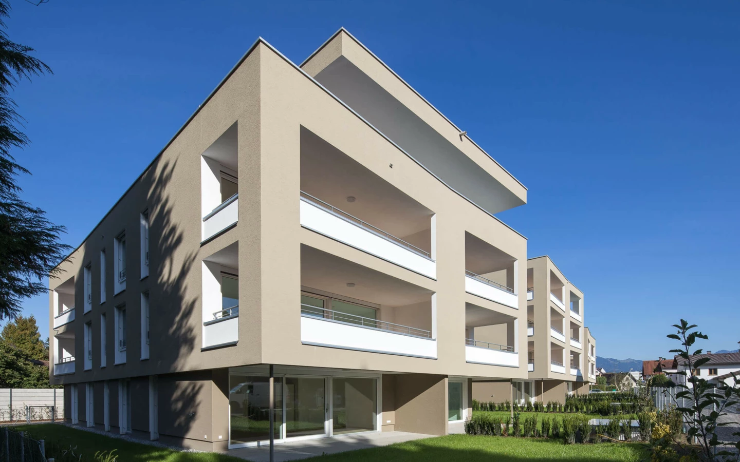 Moderne Wohnanlage Widum in Lustenau mit beigefarbenen Fassaden, mehreren Balkonen und grünen Rasenflächen unter blauem Himmel.