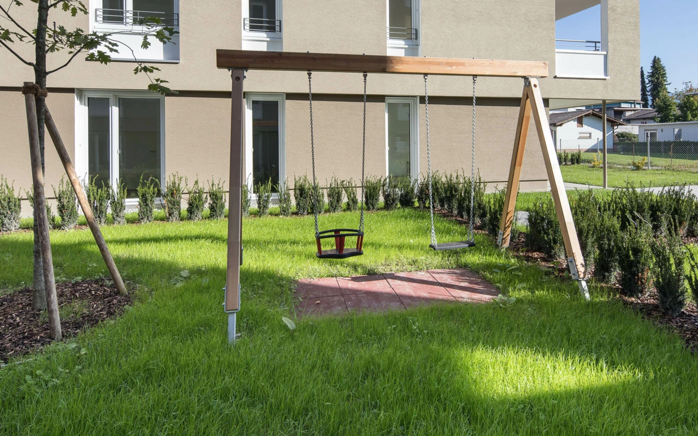 Ein Kinderspielplatz mit Schaukeln auf einer grünen Wiese vor einem modernen Wohngebäude der Wohnanlage Widum Lustenau.