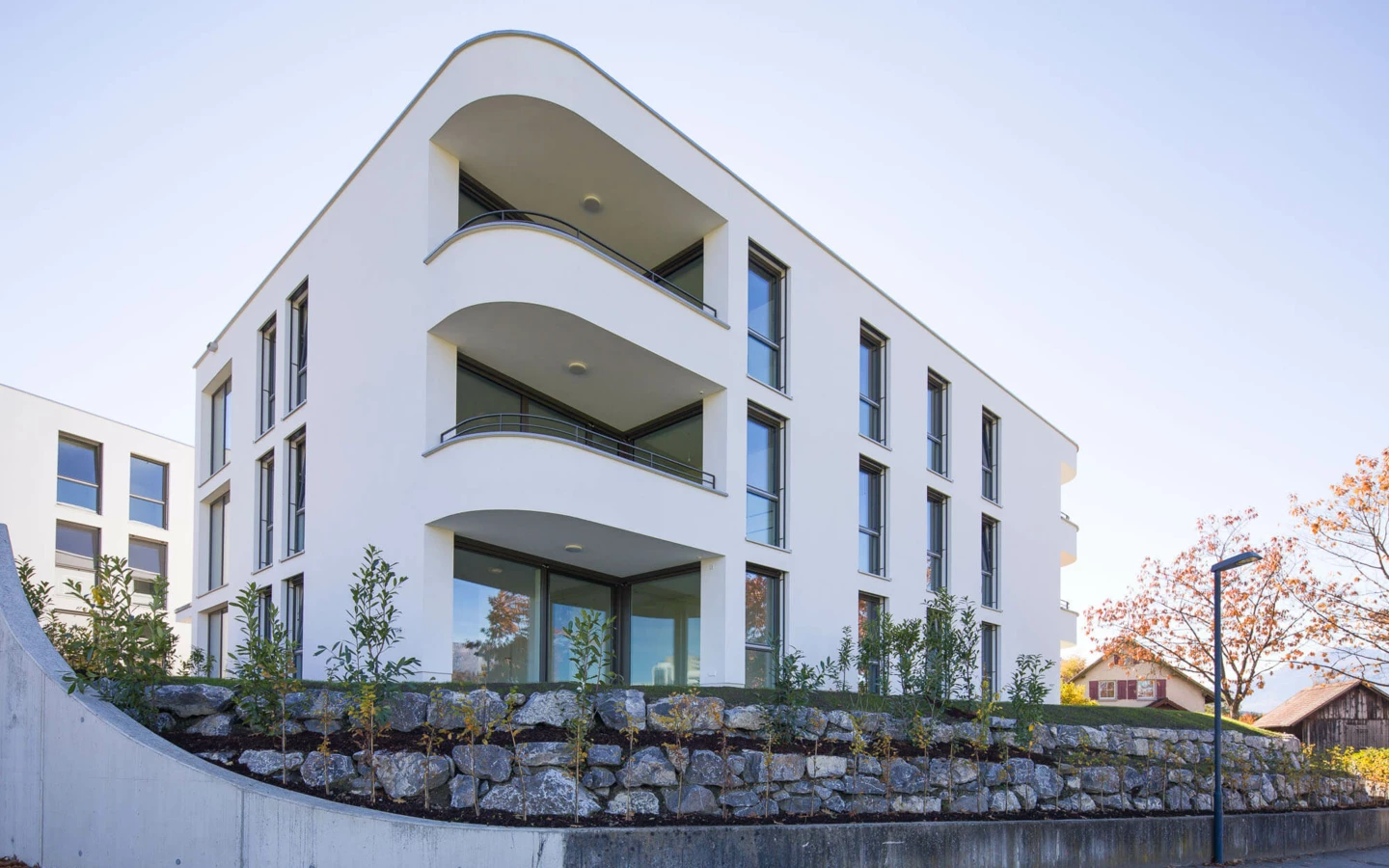 Modernes, weisses Wohngebäude mit geschwungenen Balkonen und grossen Fenstern, eingebettet in eine begrünte Steinmaueranlage (Wolf-Huber-Straße, Wohnanlage, Feldkirch).