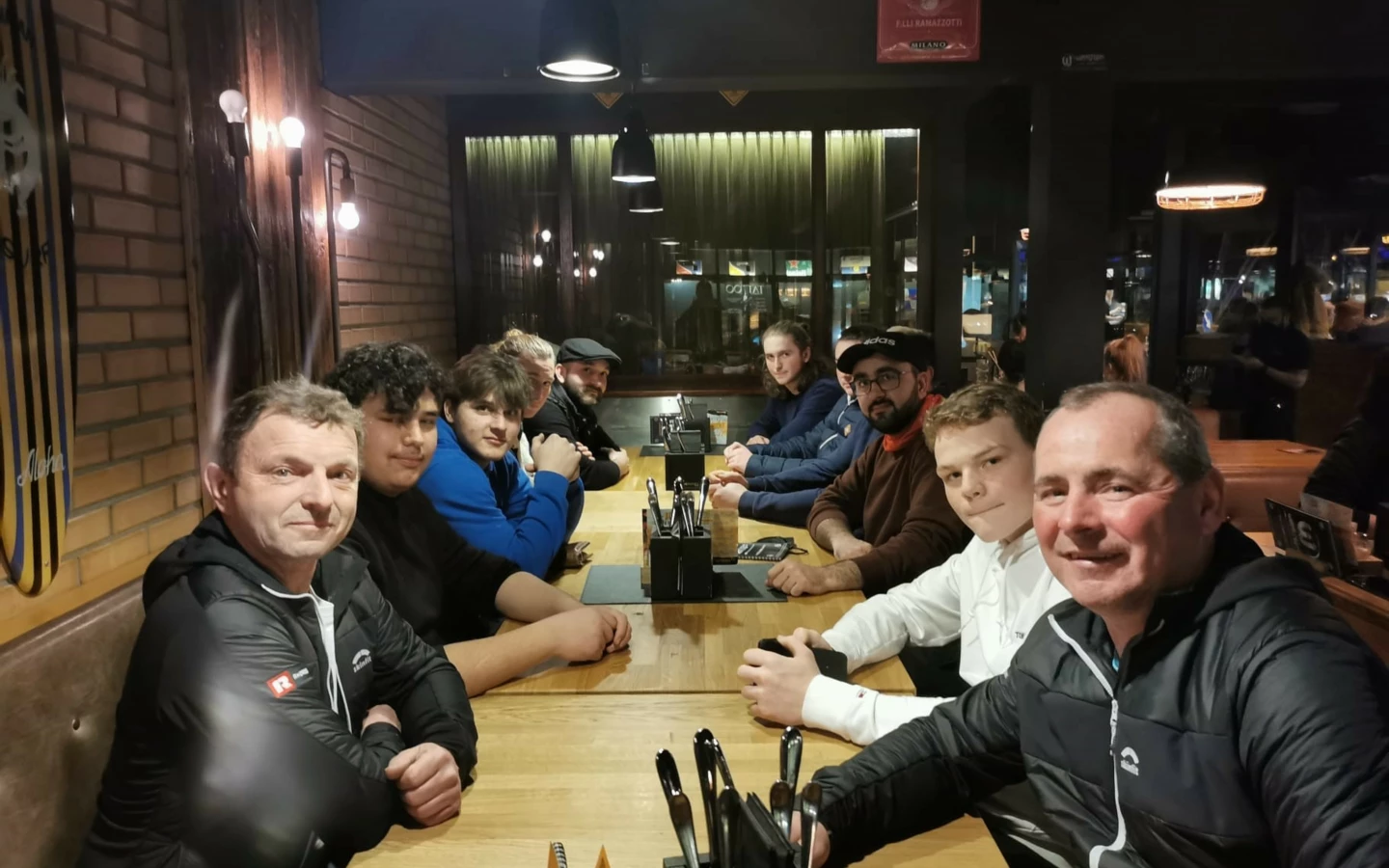 Eine Gruppe junger Männer und zwei ältere Männer sitzen gemeinsam an einem langen Holztisch in einem Restaurant und schauen in die Kamera (Lehrlingsabend).