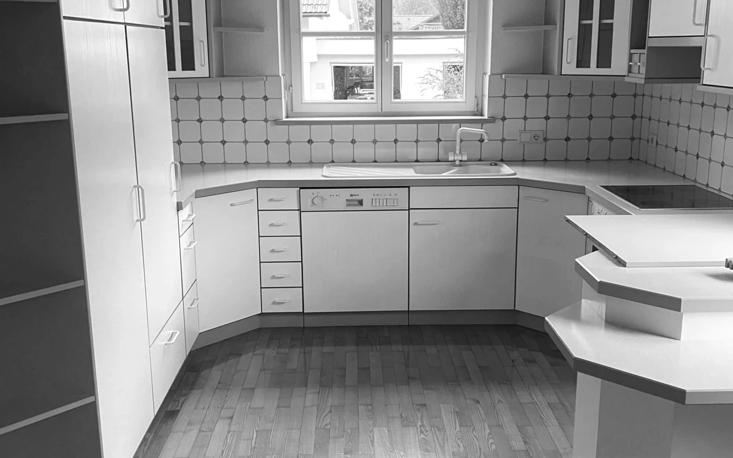 Eine leere, unrenovierte Küche mit weißen Schränken, einer Spüle, einem Geschirrspüler und Holzfußboden, aufgenommen vor der Generalsanierung Rhomberg Bau EFH Bohle.