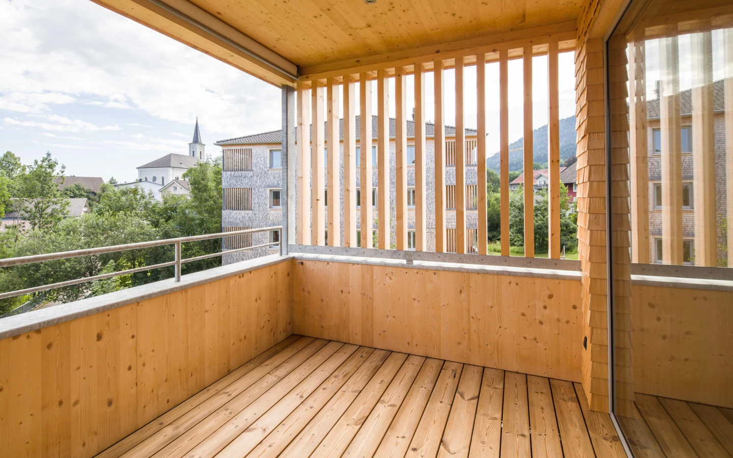 Blick von einem holzverkleideten Balkon mit Holzlatten auf eine Wohnanlage in Alberschwende und einen Kirchturm