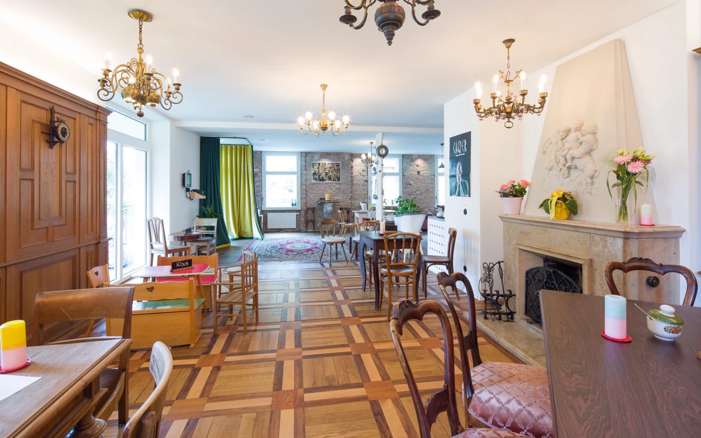 Ein geräumiges Interieur mit einem Fischgrätparkettboden, Kronleuchtern, Tischen und Stühlen, einem hellen Kamin und großen Fenstern mit Vorhängen (Wunderkind, Backstube).