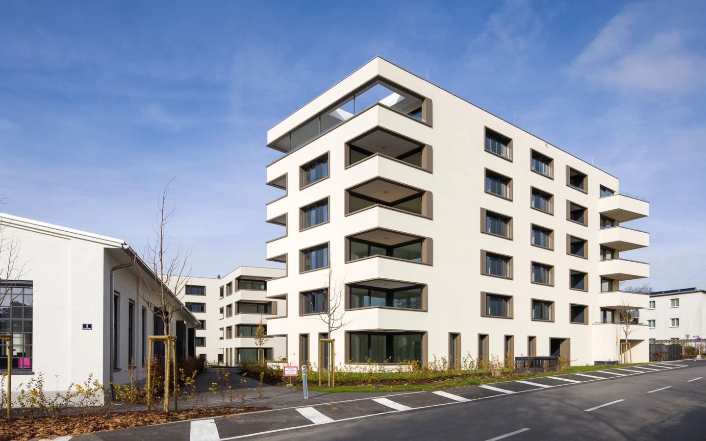 Moderne, weiße Mehrfamilienhäuser mit Balkonen und großen Fenstern unter blauem Himmel, mit einer Straße im Vordergrund (Bäumleareal).