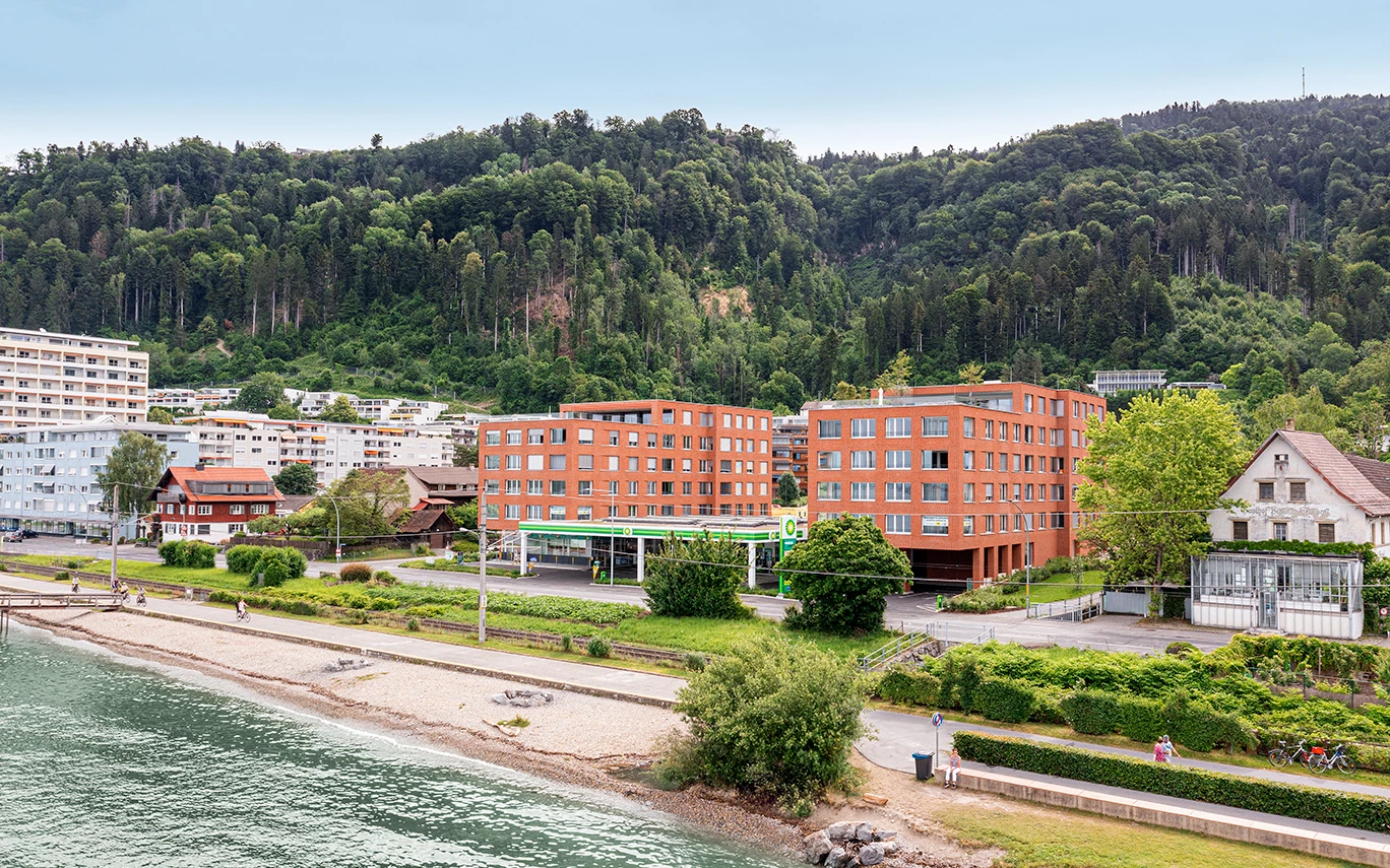 Zwei moderne Gebäude am Ufer, dahinter weitere Gebäude und ein bewaldeter Hügel mit Blick auf den Bodensee (Seeblick, Lochau).