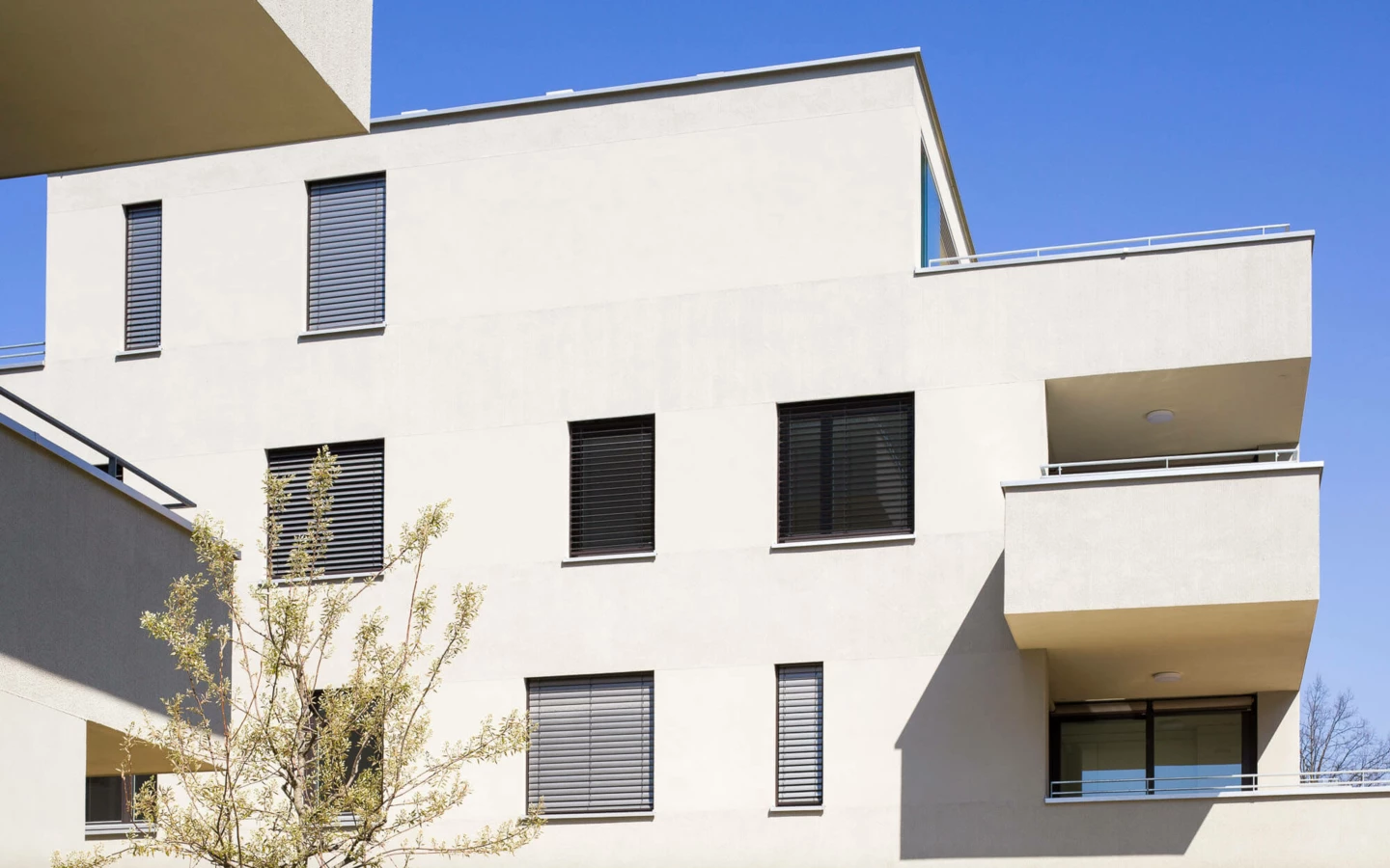Moderne Wohnanlage mit beiger Fassade, Balkonen und geschlossenen Jalousien unter blauem Himmel (Wuhrbaumweg)