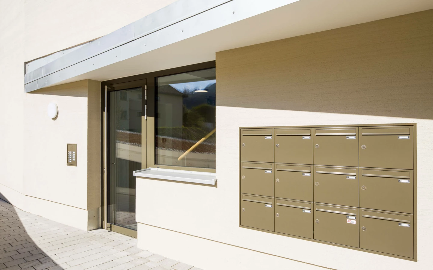 Eingangsbereich der Wohnanlage Feldgasse Dornbirn mit Briefkästen und Gegensprechanlage. Die Fassade ist hellbeige, und der Boden ist mit hellen Pflastersteinen ausgelegt.