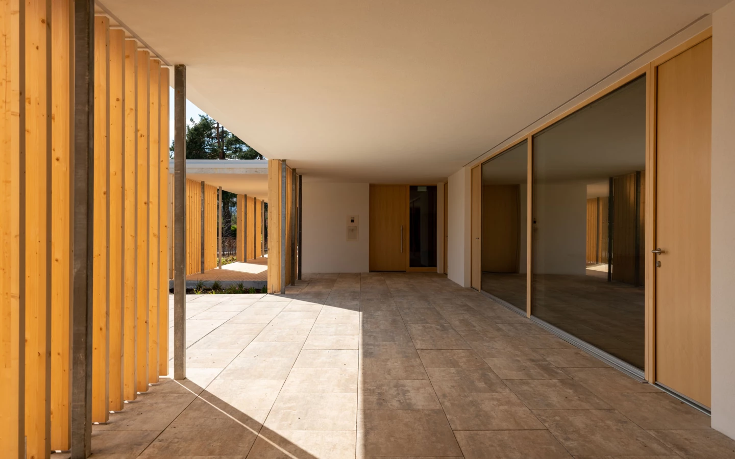 Moderner Eingangsbereich mit hellbraunen Fliesen, Holztüren und einer Reihe vertikaler Holzlamellen, teilweise im Schatten (Wolfurt, Dieter).