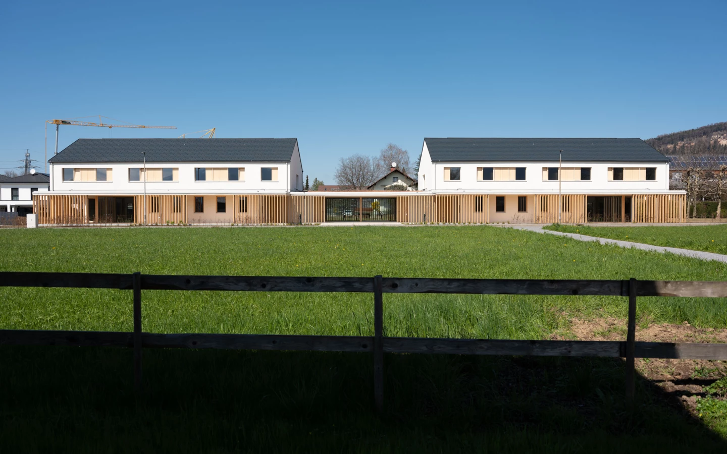 Zwei moderne, zweistöckige Gebäude mit Holzfassade und dunklem Dach, von grünen Wiesen und einem Holzzaun umgeben (Wolfurt, Dieter).
