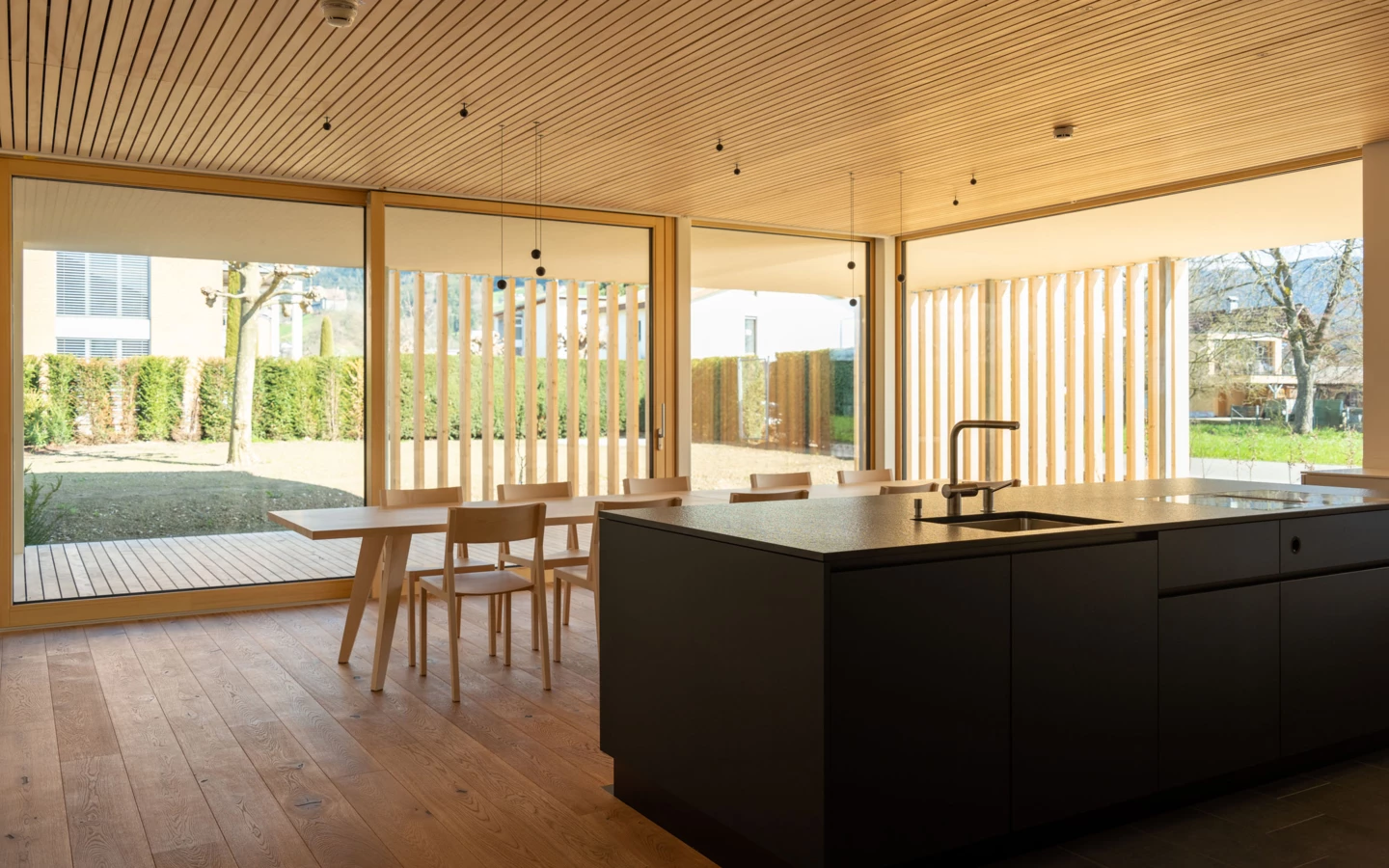 Moderne Küche mit dunkler Kochinsel und Spüle, einem Holztisch mit Stühlen und großen Fenstern zum Gartenbereich (Wolfurt, Dieter).