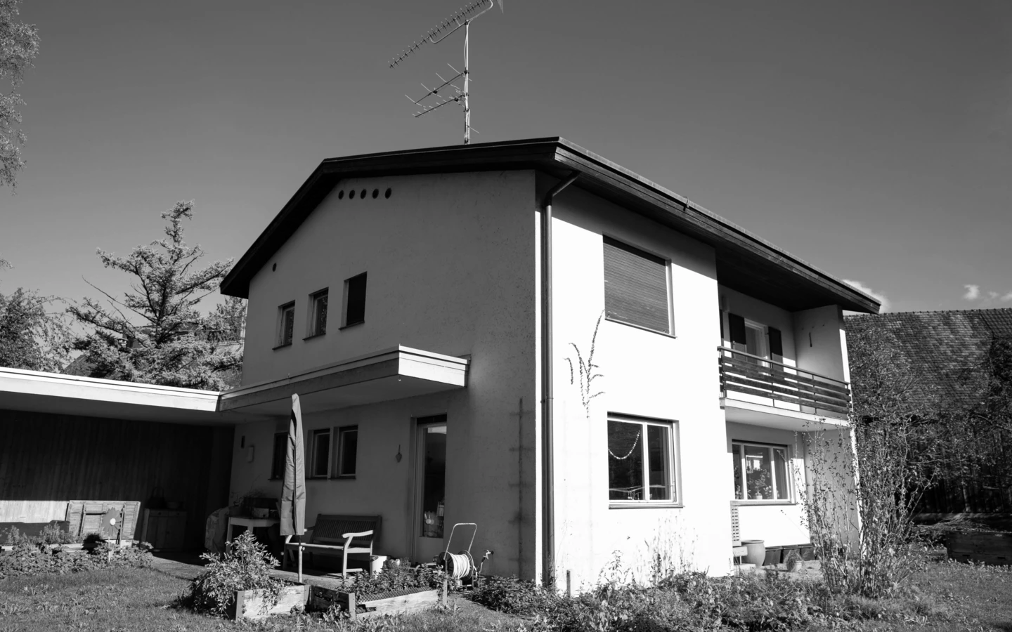 Schwarz-Weiß-Ansicht eines Einfamilienhauses in Dornbirn mit Antenne auf dem Dach, einem Carport und Gartenbereich (Generalsanierung, Holzoptik).