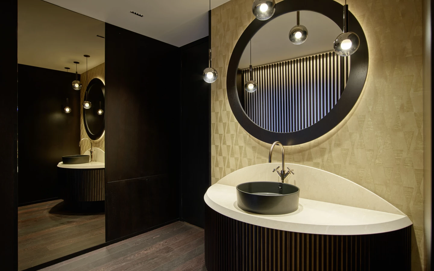 Modernes Badezimmer mit ovalem Spiegel, runder Waschtischplatte und goldener Tapete, beleuchtet von Pendelleuchten (Huber, Rathausstr).