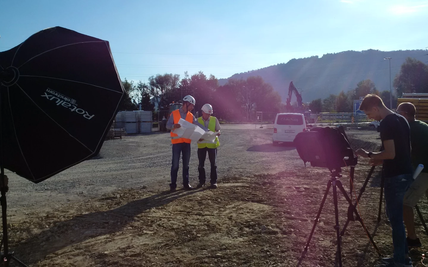 Zwei Bauarbeiter in Warnwesten und Helmen prüfen Baupläne auf einer Baustelle, während ein Fotograf filmt.