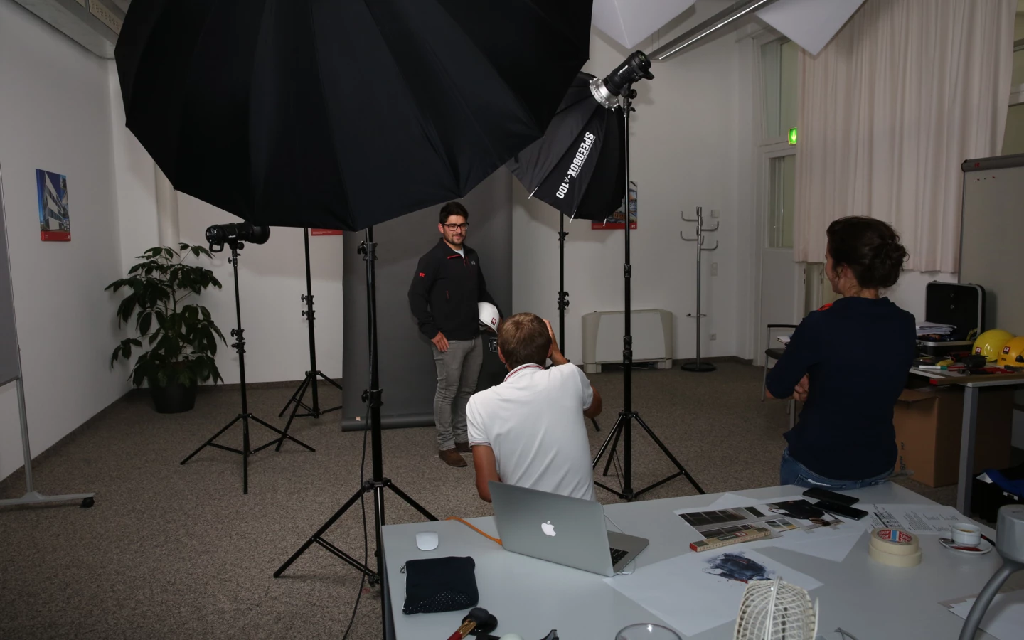 Ein Mann posiert für ein Fotoshooting mit Blitzgeräten und Regenschirmen, während ein Fotograf und ein anderer Mann zusehen.