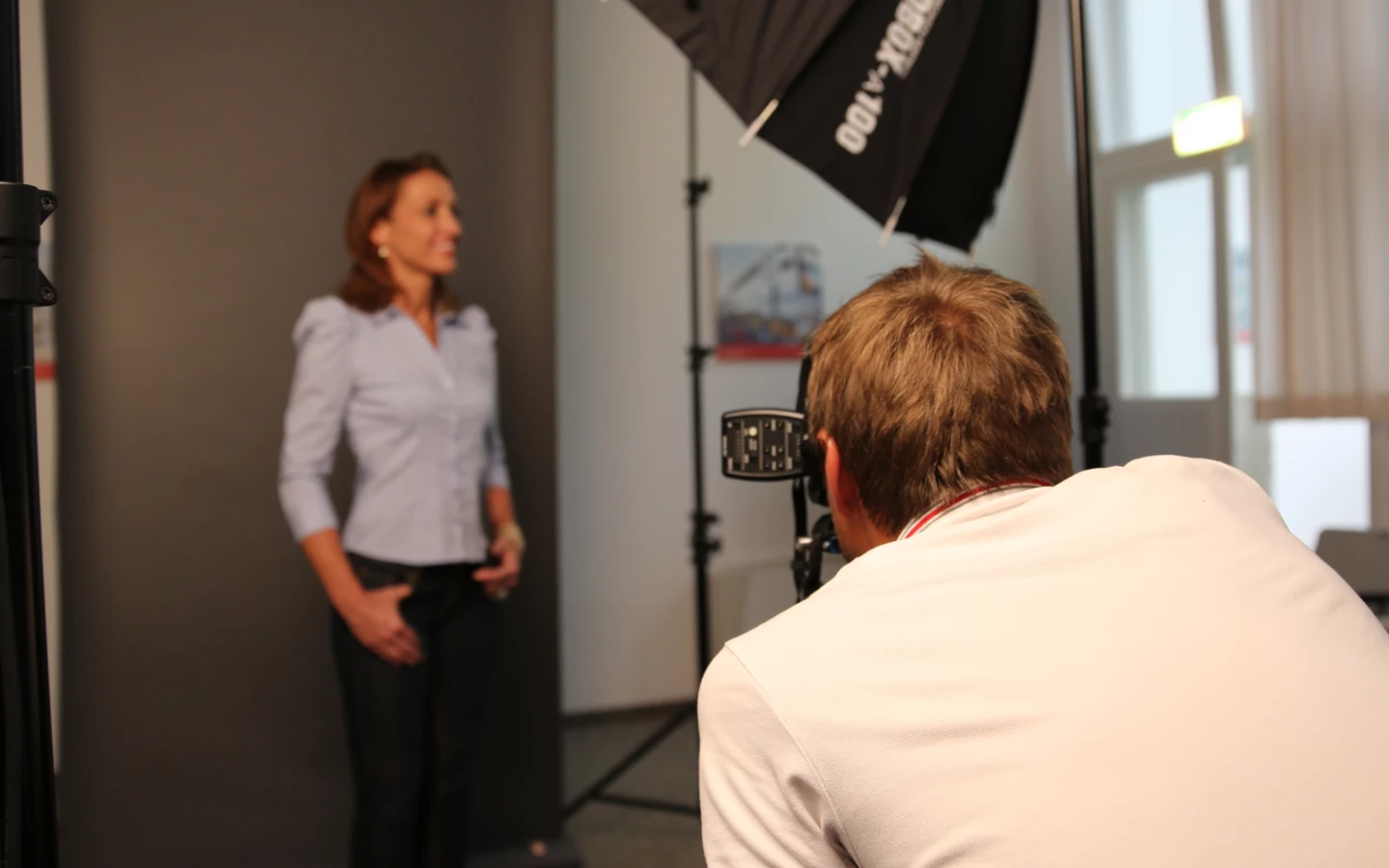 Ein Fotograf macht Aufnahmen einer Frau mit einer Kamera, umgeben von professioneller Beleuchtung und einem grauen Hintergrund.